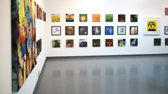 A photograph of the exhibition showing rows of paintings inspired by record covers and one larger portrait of Phil Collins in bold black pen on a yellow background. On the right side you can see a large painting of a rock concert from the side.
