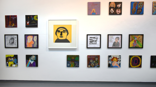 A photograph of the exhibition showing rows of paintings inspired by record covers and one larger portrait of Phil Collins in bold black pen on a yellow background.