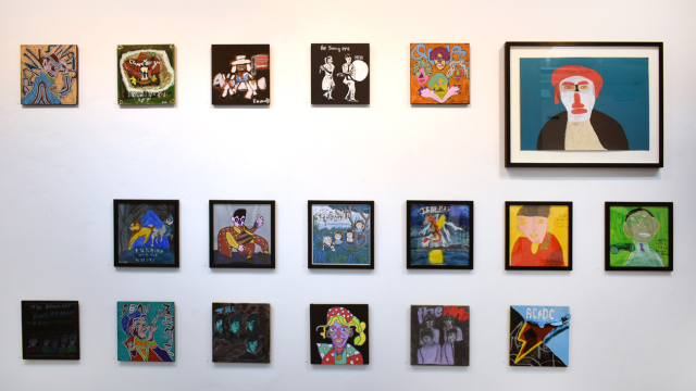 A photograph of the exhibition showing rows of paintings inspired by record covers and one larger portrait of Elton John painted in teal, red and white colours.