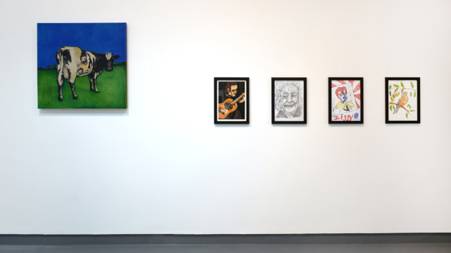 A photograph of the exhibition showing a large square painting of a cow on a vivid azure sky and emerald green grass. Alongside four smaller framed works including a pencil drawing of Noddy Holder, and a watercolour and ink depiction of a chaffinch on a branch