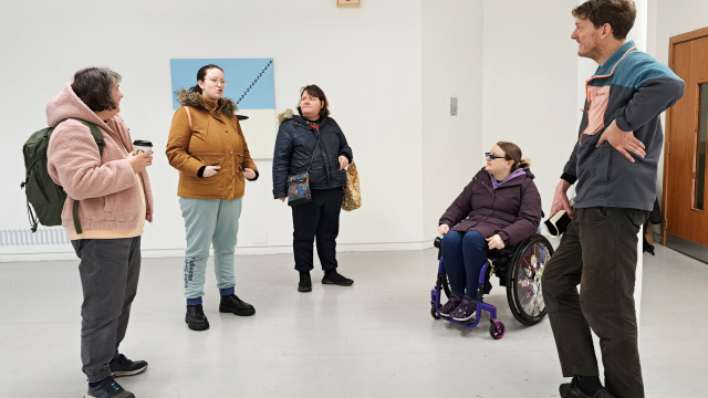 A group of people who are dressed up for the cold and rain are in a semi circle inside of a white walled gallery.