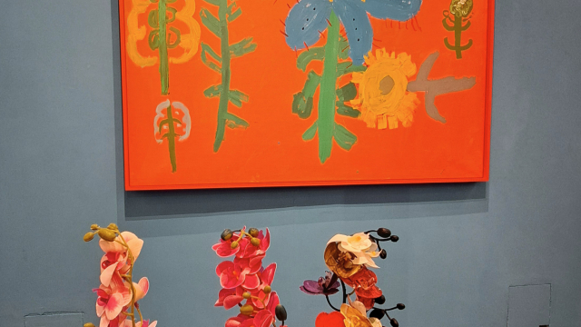 An exhibition space with blue walls. There is an art piece of flowers hanging from the wall. Closer to the camera, there is a table with three colourful geranium plants and some artistic crokery.