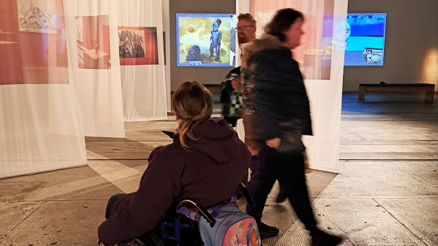 A wheelchair user and two other people in a gallery space with long white pieces of fabric hanging from the ceiling. There are two screens in the wall at the end of the room.