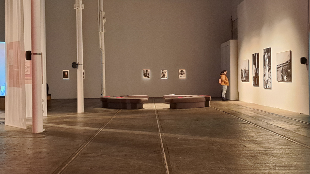 A big spacious gallery with white walls and pillars. There are different clusters of art on the walls with spotlights on them. There is a person wearing a yellow jacket staring at a piece.