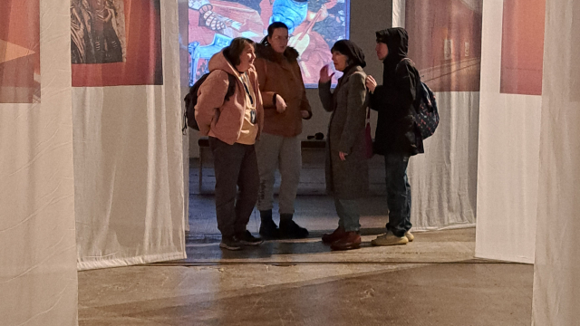 Four people gather to talk between white sheets in a gallery space. The sheets are long and hanging from the ceiling in a pattern. There is a screen with an artwork at the very end of the room.