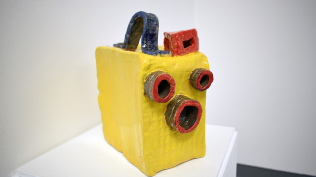 ceramic camera A bright yellow ceramic sculpture of a box brownie film camera. It has three brown ring that are the lenses on the front. A boxy red viewfinder on the top and a curved blue strap at the top just big enough for your hand.