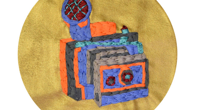 On a metallic gold fabric background is a thickly textured embroidery of an accordian camera, in blocky colours of brown, orange and blue, with added detail around the buttons, flash and lense in a metallic dark red and a turquoise blue.