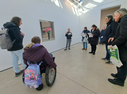 The walking group standing round a woman reading from a sheet of paper. They are in a large white gallery with a large artwork on the wall showing a tall vase with large pink flowers.