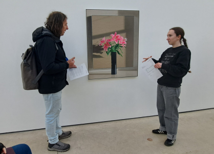 A member of the walking group and Mae stand either side of one of the large flower paintings from the exhibition. The painting has a brown canvas background with a tall black vase and large pink blooming flowers with green leaves.