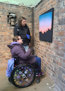 two members of the walking group are looking at an artwork hanging on a bare brick wall. One person is standing, the other is in a wheelchair. The artwork is on a large black background and looks 3d. It is square shape at the top and has a broken ragged edge at the bottom. It starts as bright blue at the top which blends into a lighter blue which blends in to a light purple/ grey colour before finally blending into a light pink at the bottom.