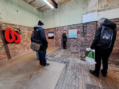 a photograph of an open gallery with righ wooden floor boards and exposed brick walls, There are 3 of people standing around and some artwork on the walls.