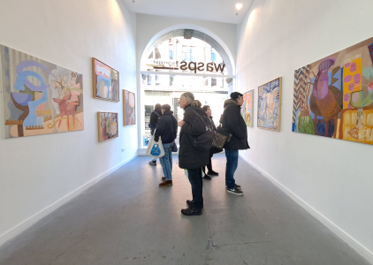 a group of people standing the BriggaiT gallery looking at various pieces of abstract art hanging either side of them on white walls.