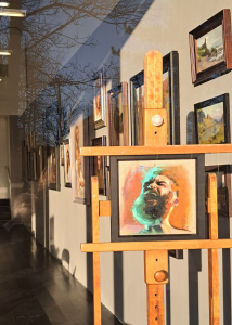 A photograph taken through a window. You can see the reflection of trees in the left hand corner of the window. Through the window you can see lots of brown framed paintings hanging on a gallery wall to the right. There is an easel as the front holding a black framed painting of a bearded man throwing his head back with his mouth open. The face has highlights of turquoise and it is on an orange background.