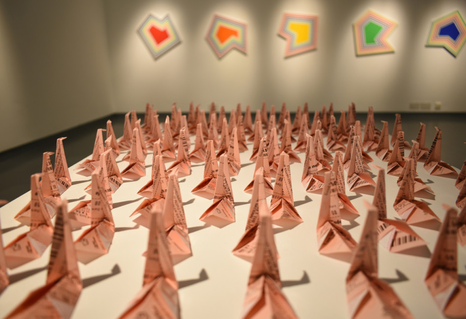 A close up photographs showing lots of light pink coloured paper cranes on a plinth, made of lottery tickets. In the background are 5 multi coloured bright abstract shapes