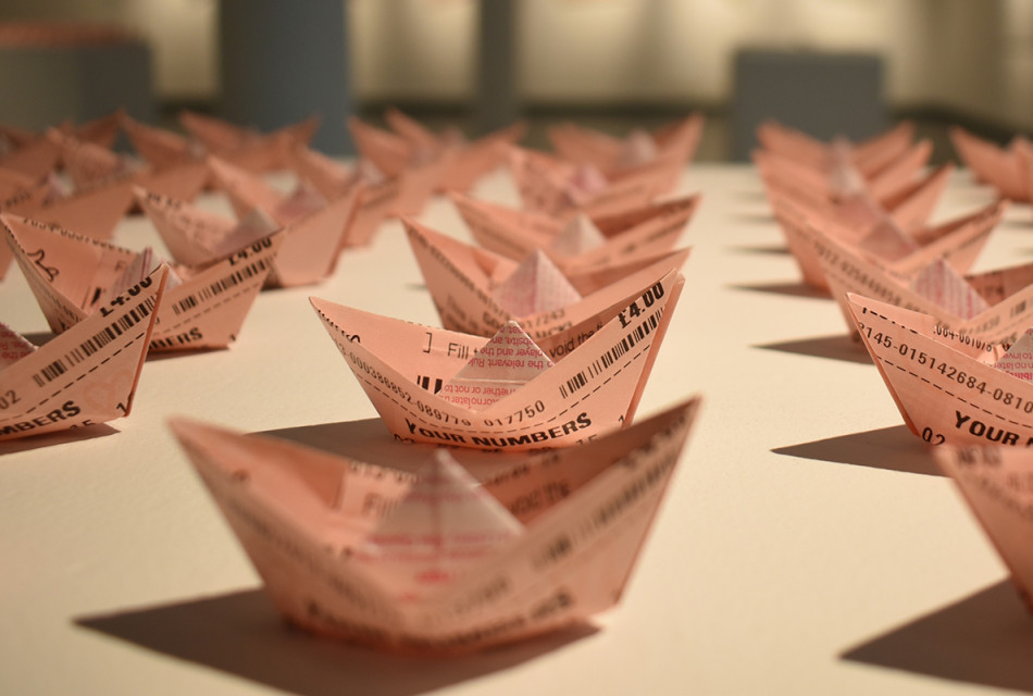 A very close up photograph showing lots of light pink coloured boats, made of lottery tickets. You can see the different sides of the paper showing through the origami creases.