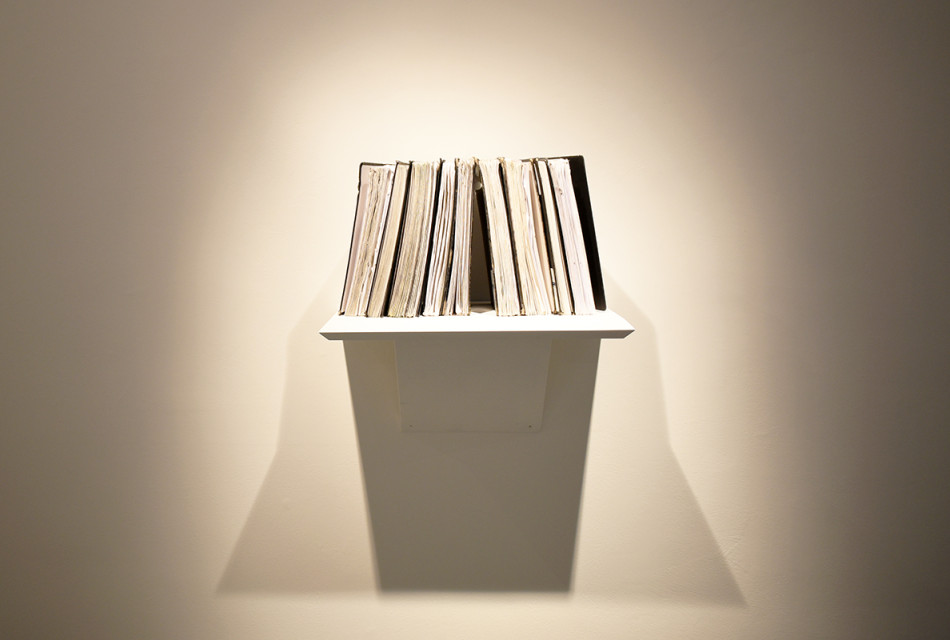 Photograph showing a small white shelf on a white gallery wall holding lots of sketchbooks propped up on it, with the pages facing outward and the spines towards the wall.