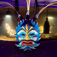 Colorful blue mythical mask with curved horns, black feathers, gold details, and a red mouth hangs in a dim, mysterious exhibit.