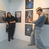 Two visitors explore a contemporary gallery lined with framed portraits; a man photographs a woman smiling in front of the gallery wall.