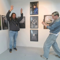 Two men pose playfully in a contemporary photo gallery, surrounded by framed portraits; one raises his hands while the other photographs him.