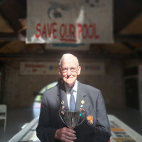 Smiling elderly man in a dark suit holds a trophy beneath a “Save Our Pool” sign
