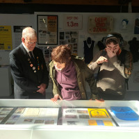 At a museum exhibit, an elderly man, a woman and a young man look at documents in a display case, with memorabilia and signs behind them.