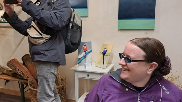 A man in a grey jacket takes a photo with his phone while a smiling woman in a purple jacket sits in a wheelchair in an art gallery setting.