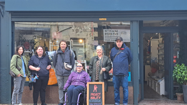 A group of six people stand outside the originalartists shop, with one person in a wheelchair next to a sign for a Project Ability art exhibition.