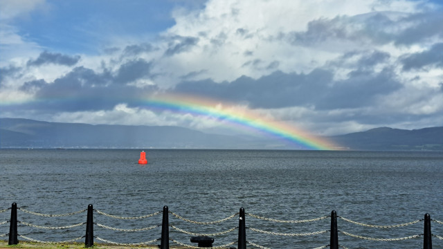 A vibrant rainbow arches over a calm, dark sea with a bright red buoy, framed by a black metal fence and a brick walkway under a cloudy sky.